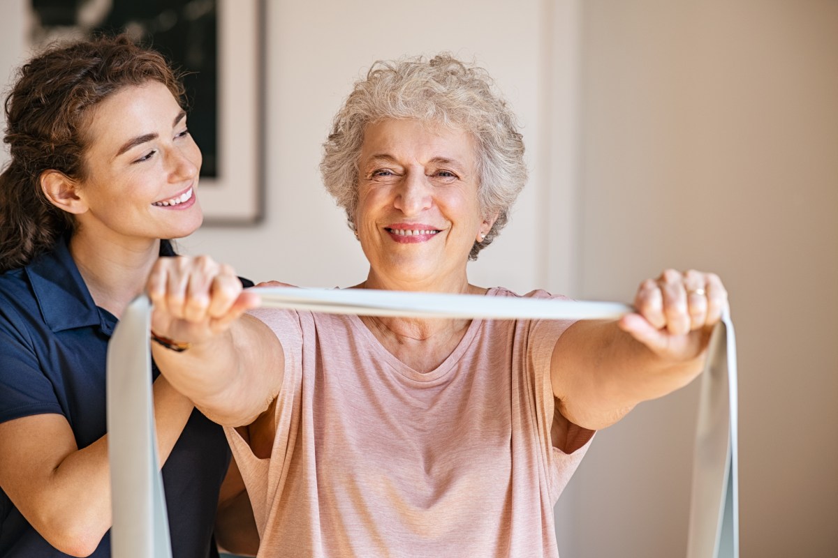 Young female trainer assisting senior woman with resistance band. Rehabilitation physiotherapy worker helping old patient at nursing home. Old smiling woman with stretch band being coached by personal trainer or physiotherapist in clinic.